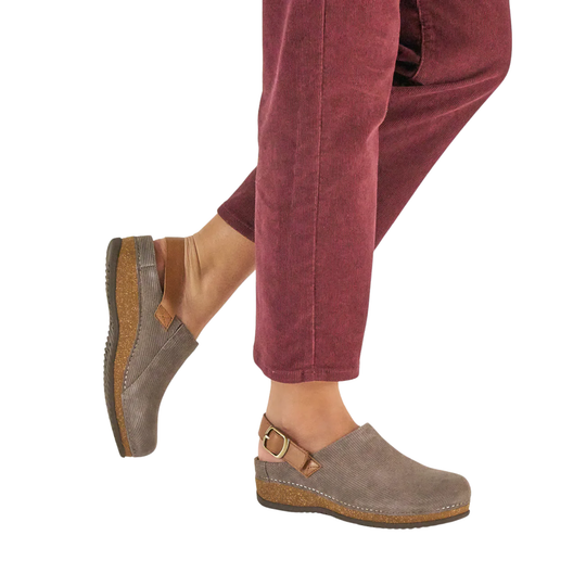 A person wearing taupe suede clogs with cork soles and a small buckle strap, paired with rust-colored pants, shown against a plain white background.