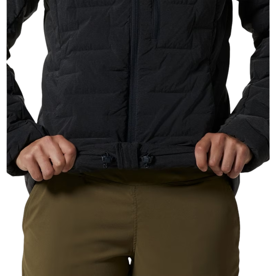 Close-up of a person wearing a black quilted jacket and olive green pants, pulling the jacket hem down with both hands against a white background.