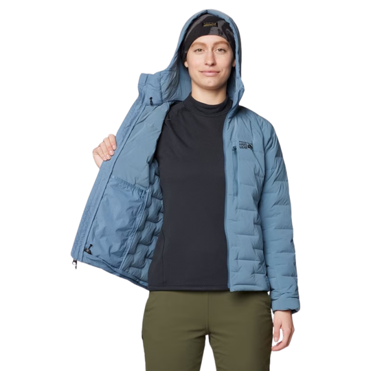 A woman wearing a light blue hooded puffer jacket over a black shirt and olive green pants, holding the jacket open against a plain white background.