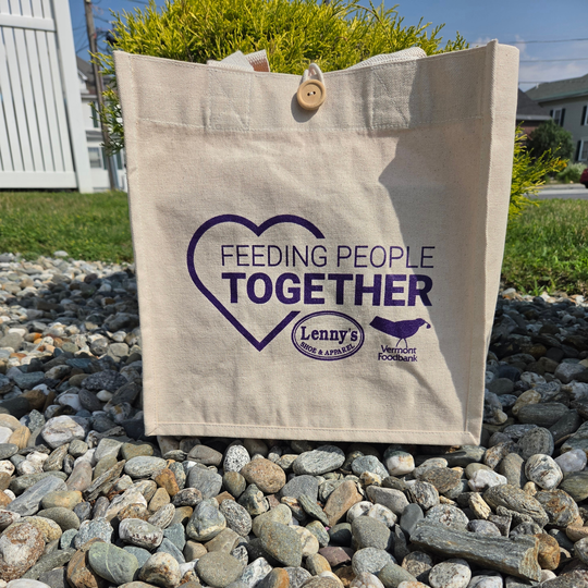 A beige canvas tote bag with a wooden button closure stands on a bed of small gray pebbles outdoors. The bag features a purple printed heart and the text 'FEEDING PEOPLE TOGETHER' along with the 'Lenny's Food & Apparel' logo.