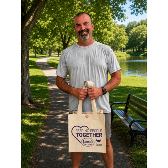 A smiling man standing on a park path holding a cream canvas tote bag printed with a purple heart and the words 'FEEDING PEOPLE TOGETHER' and 'Lenny's Animal Rescue'.