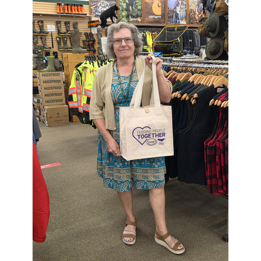 An older woman stands inside a retail store holding a cream canvas tote bag with a purple heart graphic and the words 'Together People United'. The store background features clothing racks, boots, and other merchandise.