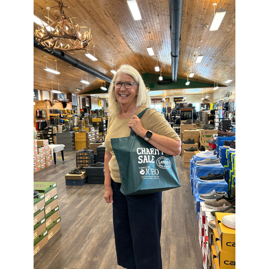 A smiling blonde woman wearing glasses stands inside a retail store holding a teal tote bag printed with 'CHARITY SALE' and the JCEO brand logo. The store has a wood ceiling, chandeliers, and shelves of products.