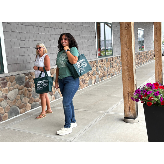 Two women standing on a covered walkway outside a building, each carrying a dark green Charity Sale 2023 tote bag with white text.