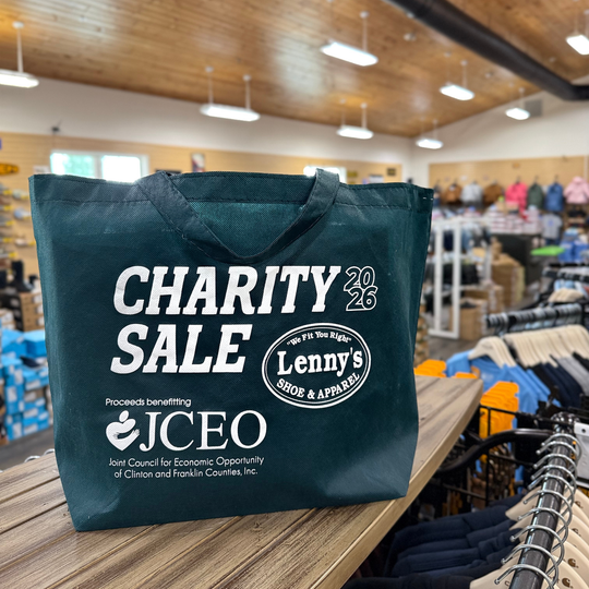 A dark teal reusable shopping bag printed with 'CHARITY SALE 2026' and 'Proceeds benefiting JCEO' in white text, placed on a wooden counter inside a retail store with clothing racks and ceiling lights blurred in the background.