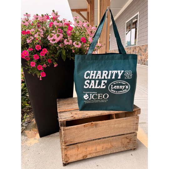 A dark teal tote bag with white printed text reading 'CHARITY SALE' and 'Lenny's' sits on a rustic wooden crate outdoors next to a large black planter filled with pink flowers.