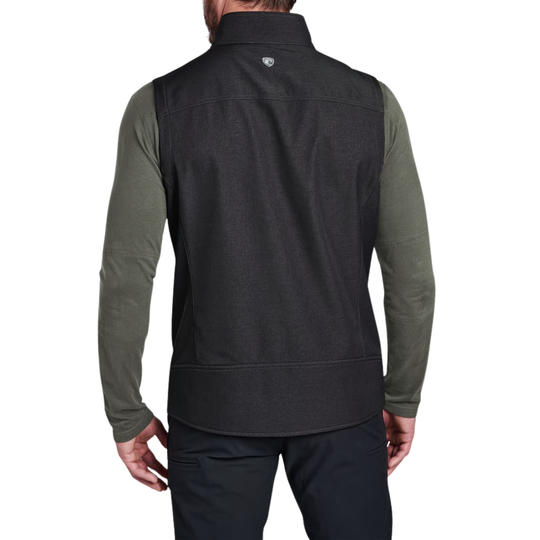 Rear view of a man wearing a dark charcoal gray sleeveless zip vest over an olive green long-sleeve shirt and dark pants, against a plain white background.