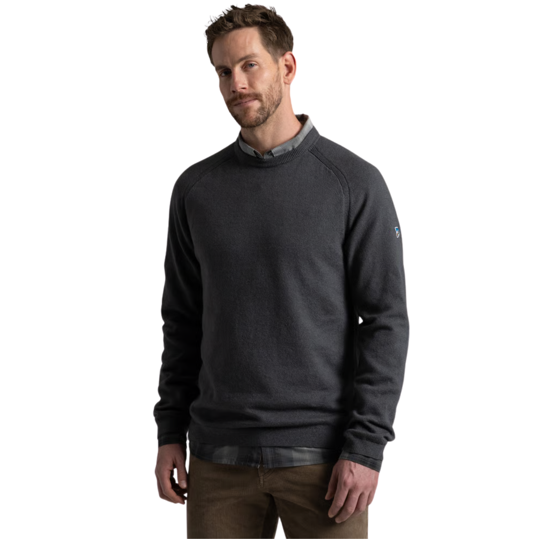A man wearing a dark charcoal gray long-sleeve crewneck sweater over a collared shirt, paired with brown pants, photographed against a solid white background.