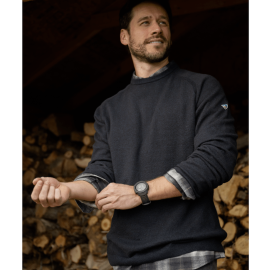A man wearing a dark charcoal crewneck sweater with a small white logo on the left sleeve, standing in front of a stack of firewood.