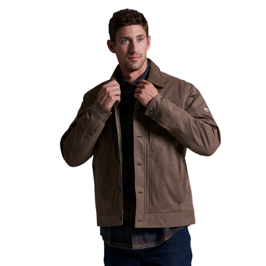 A man wearing a brown utility jacket over a dark shirt and blue jeans, standing against a solid white background.