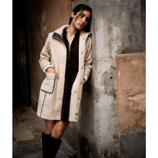 A woman leans against a textured stone wall wearing a cream-colored shearling-style coat over a dark dress and black knee-high boots.
