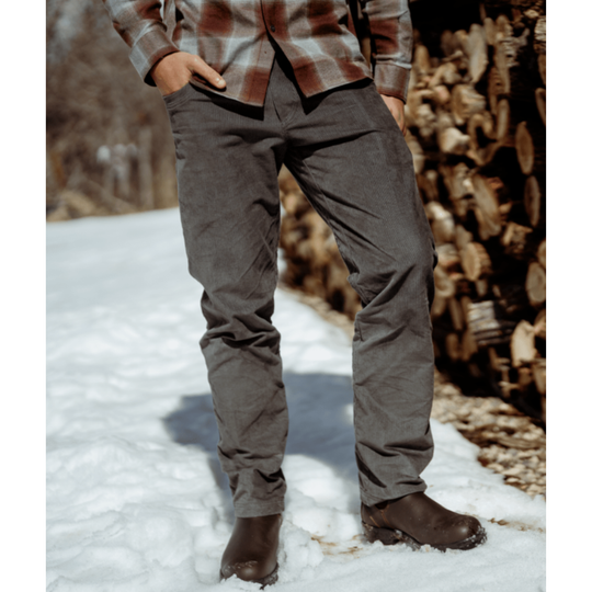 A person standing outdoors in snow next to a stack of firewood, wearing dark gray pants, a brown and beige plaid shirt, and dark brown boots.