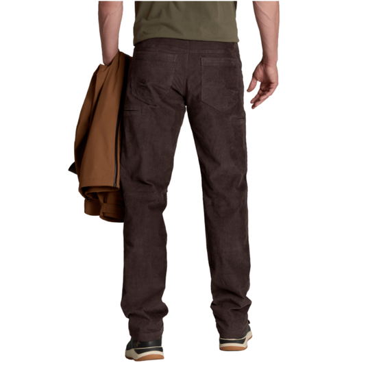 Rear view of a person wearing dark brown work pants, holding a brown jacket over one arm against a plain white background.