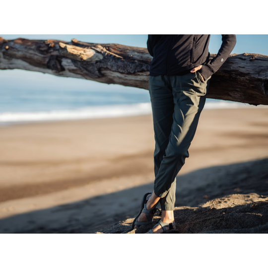 A person standing on a sandy beach leaning against a large fallen log, wearing dark olive green pants, a black long-sleeve top, and sandals.