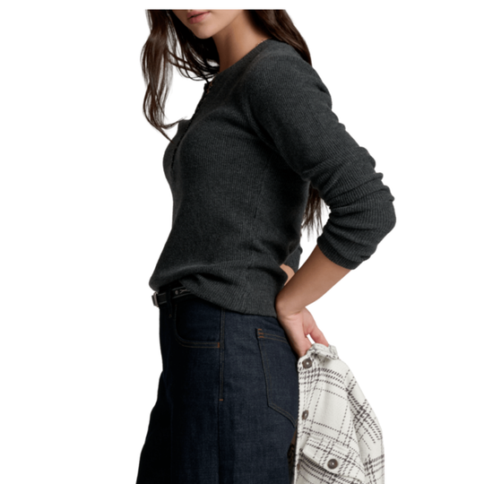 A woman wearing a dark charcoal gray ribbed knit sweater and dark blue jeans, holding a white and gray plaid shirt in her hand against a plain white background.