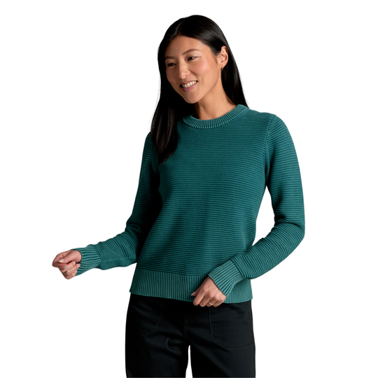 A woman wearing a dark teal-green ribbed knit crewneck sweater and black pants, standing against a plain white background.