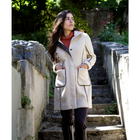 A woman wearing a cream-colored textured coat with black piping, a rust-brown turtleneck, and black pants, standing on stone stairs outdoors.