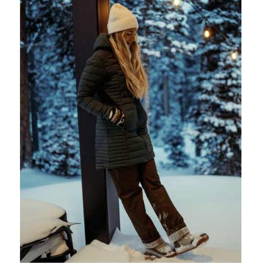 A woman wearing a dark olive green quilted puffer jacket, brown snow pants, a white knit beanie, and gloves stands in a snowy outdoor setting at dusk.