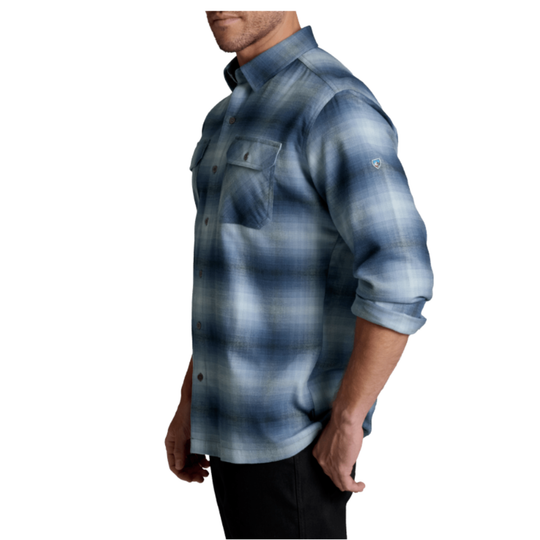 A man wearing a blue and gray plaid button-down shirt with rolled-up sleeves, shown from a side profile against a white background.