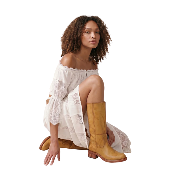 A model with curly brown hair is kneeling on a white studio background, wearing a white off-the-shoulder lace dress and tan leather knee-high boots.