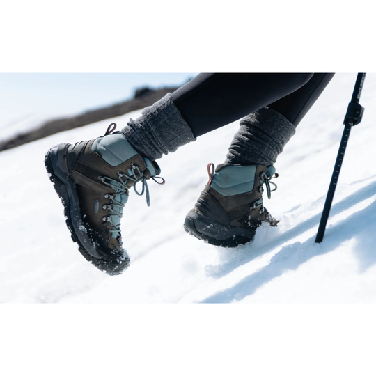 A close-up of a person's legs in black leggings and gray knit socks, wearing brown and gray hiking boots with snow on them, while walking in the snow. A ski pole is visible on the right.