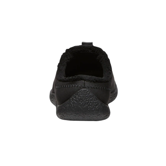 A single black slip-on shoe shown from the back, featuring a textured rubber sole and a soft, fuzzy black lining, set against a plain white background.