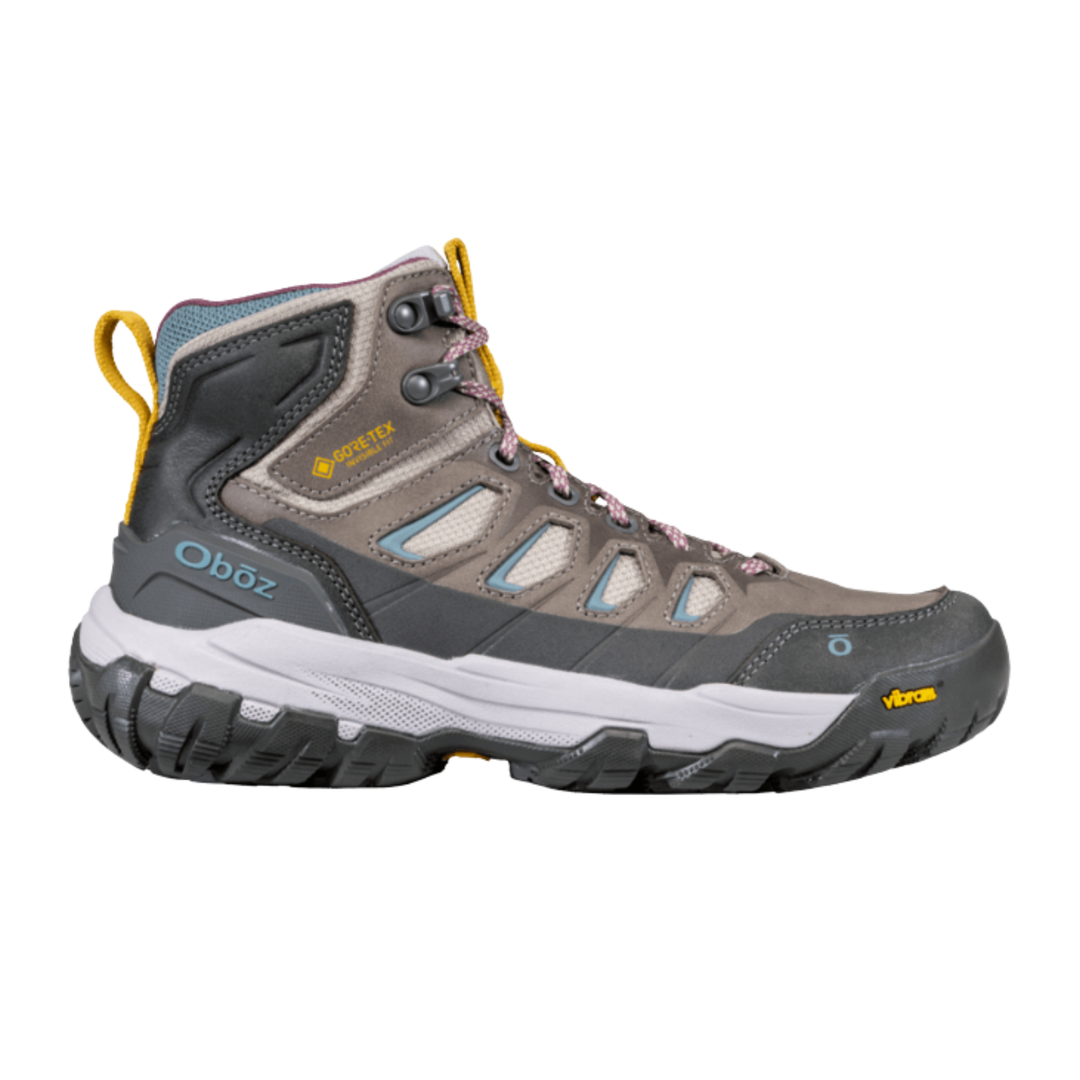 A mid-cut hiking boot from Oboz, featuring a gray and brown suede and synthetic upper with light blue mesh panels, a white midsole, and a black Vibram outsole.