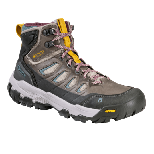 A mid-height hiking boot from oboz, featuring a gray suede and synthetic upper with black and white soles, yellow accents on the laces and toe bumper, and a GORE-TEX label on the side.