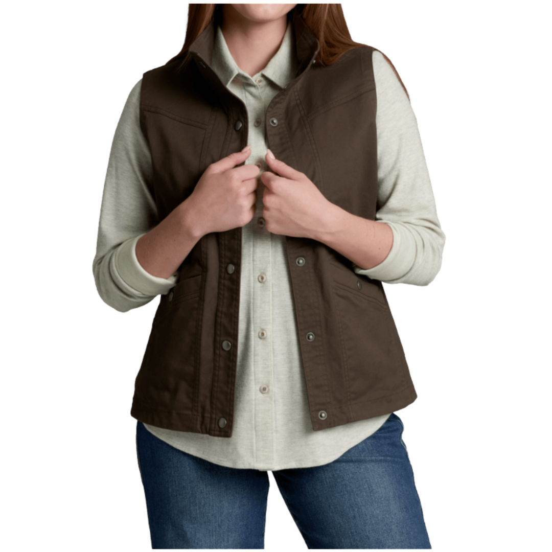 A woman wearing a brown sleeveless button-front vest over a light beige button-up shirt and blue jeans, shown from the mouth down on a plain white studio background.