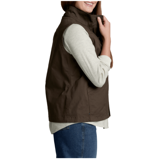 A woman wearing a dark brown sleeveless zip-front vest over a light beige long-sleeve shirt and blue jeans, shown from the side against a plain white background.