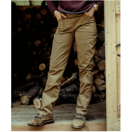 A person wearing tan work pants and brown work boots, standing on a wooden floor in front of a stack of logs. The person is cropped from the chest down, with hands in the pockets of the pants.