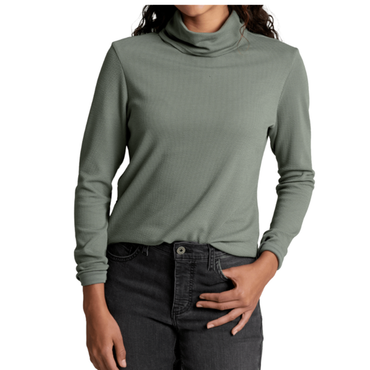 A woman wearing a sage green long-sleeve ribbed turtleneck top and dark gray jeans, shown from the neck down against a plain white background.