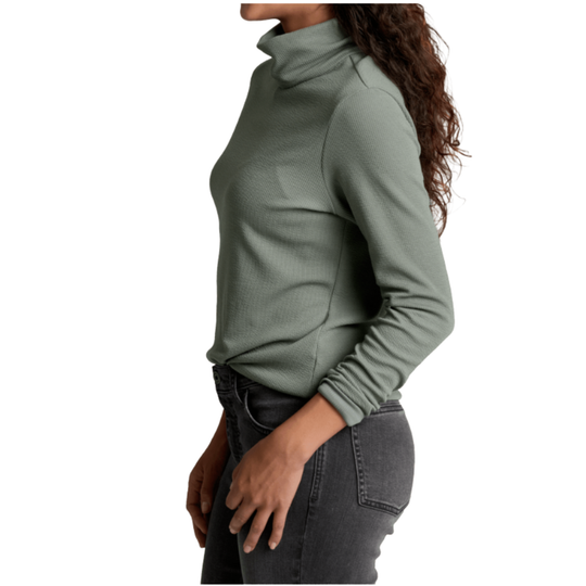 A woman shown from the side wearing a sage green long-sleeve mock neck top and dark gray jeans, against a plain white background.