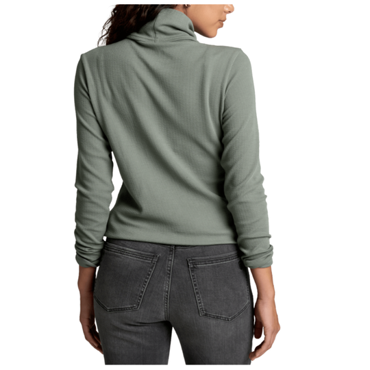 A woman shown from the back wearing a sage green long-sleeve waffle-knit turtleneck top and dark gray jeans against a plain white background.