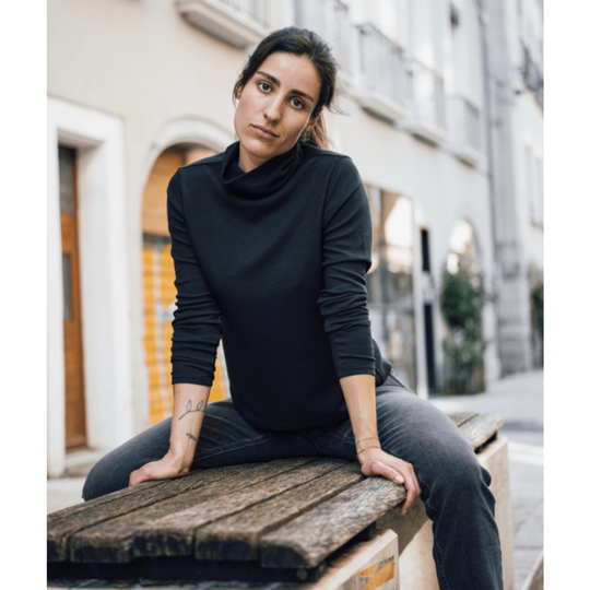 A woman wearing a black long-sleeve mock neck top and dark gray jeans sits on a weathered wooden bench in an outdoor urban setting.