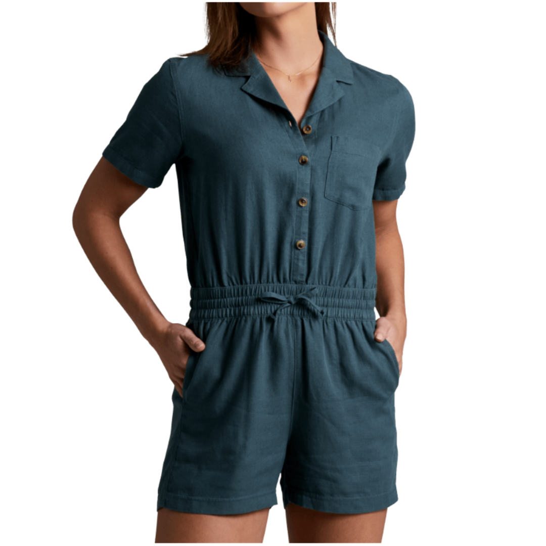 A woman wearing a short-sleeve romper in a muted teal-blue color with brown buttons, a collar, and side pockets, shown against a plain white background.