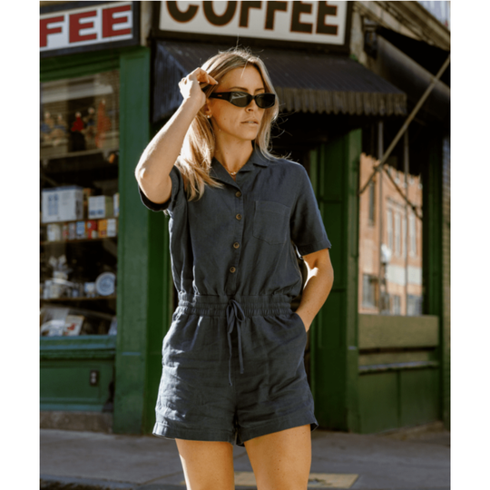 A woman wearing a dark slate blue short-sleeve romper stands on a city street in front of a green storefront with a coffee sign.