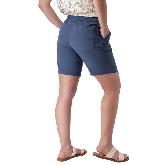 A person shown from the back wearing slate blue shorts with side pockets, paired with a patterned top and beige slide sandals, against a plain white background.