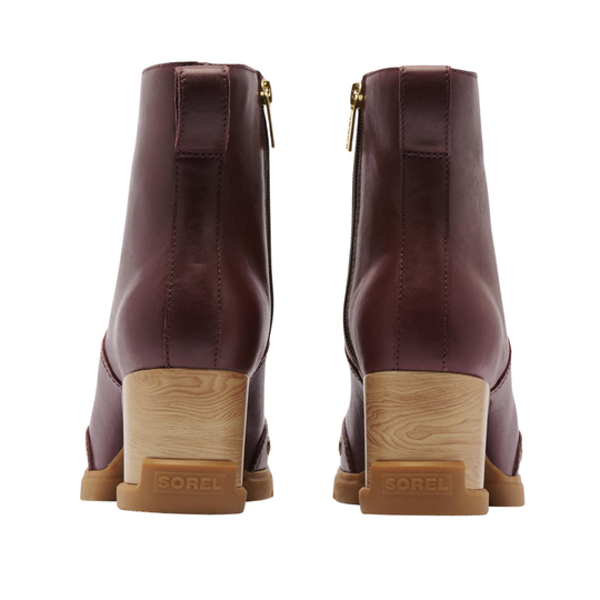 A pair of Sorel ankle boots in burgundy leather, shown from the back with their light brown wood-grain block heels and gold-tone zippers visible against a white background.