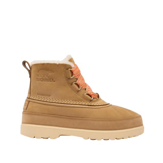 A Sorel mid-calf winter boot in tan suede with a cream shearling lining, orange braided laces, and a thick beige sole, shown from a side profile on a white background.