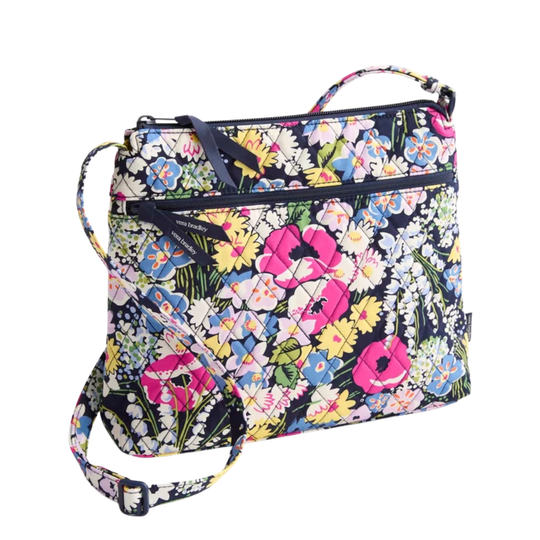 A floral print crossbody handbag with a navy blue base and a vibrant multicolor pattern of pink, yellow, blue, and white flowers. It features a top handle, a front zippered pocket, and a detachable shoulder strap.