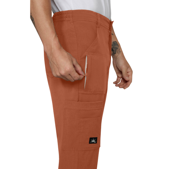 A person wearing rust-orange utility scrub pants, shown from the waist down against a white background. The model is demonstrating a side zippered pocket containing a small white tool, with a small black brand patch visible on the lower leg.