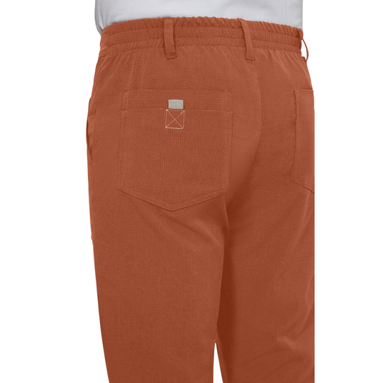 Close-up back view of a person wearing rust-orange corduroy pants with an elastic waistband, belt loops, and a back pocket featuring a small white stitched logo.