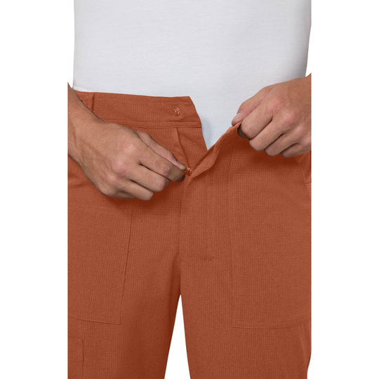 Close-up of a person wearing rust-orange corduroy trousers, pulling the waistband down with both hands over a white shirt.