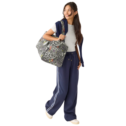 A woman modeling a navy blue track jacket and matching wide-leg sweatpants with white side stripes, carrying a large gray and white paisley print tote bag over her shoulder, against a solid white background.
