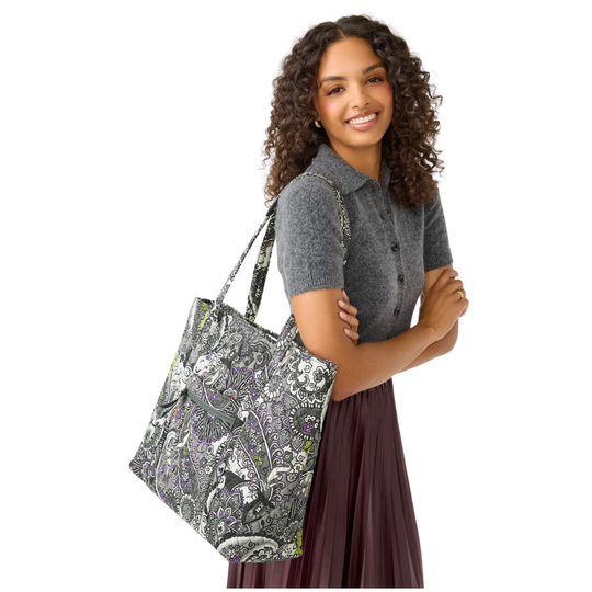 A woman with curly brown hair wearing a gray polo shirt and a dark purple skirt, carrying a large tote bag over her shoulder. The tote bag features a detailed paisley pattern in gray, white, black, and purple.