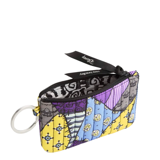 A small zippered pouch with a colorful patchwork design featuring purple, yellow, blue, and gray sections with whimsical illustrations. It has a black zipper, a black brand tag reading 'Vera Bradley', and a silver metal key ring attached on the left.