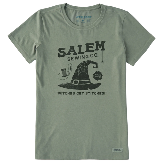 A sage green short-sleeve t-shirt with a black graphic print on the front reading 'SALEM SEWING CO.' and 'WITCHES GET STITCHES!' with a witch hat illustration.
