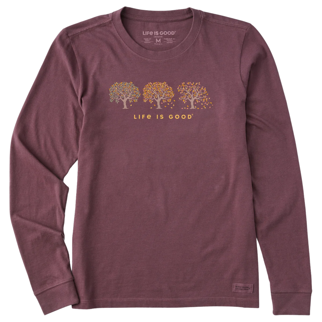 A long-sleeve crewneck t-shirt in heathered burgundy, featuring a chest graphic of three trees with orange and yellow leaves and the text 'LIFE IS GOOD' underneath. The brand 'Life is Good' is visible on the inner neck label.
