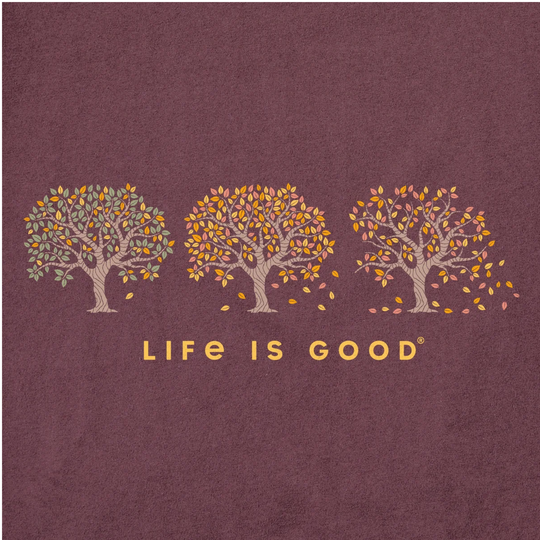 A square graphic with a textured dark mauve background, featuring three stylized trees with beige trunks and yellow, orange, and muted green leaves, above the words 'LIFE IS GOOD' in gold. A thin white vertical stripe runs along the far left edge.